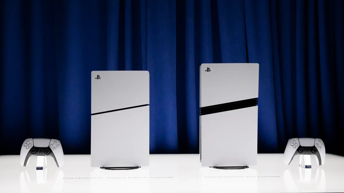 Anunciada la fecha de lanzamiento, características y precio de la PlayStation 5 Pro Dos consolas PlayStation 5 de Sony, en sus versiones digital (izquierda) y con lector de discos (derecha). Ambas consolas tienen un diseño vertical con un acabado en color blanco y una banda diagonal negra en el centro. Frente a cada consola, se puede ver un controlador DualSense de color blanco con detalles en negro. El fondo es una cortina azul, y las consolas están expuestas sobre una superficie blanca iluminada.