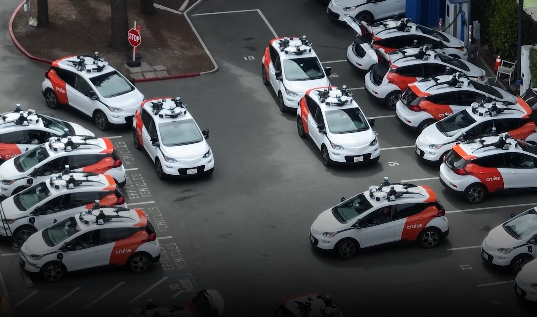 Los Robotaxis ya están aquí y la opinión pública está dividida Una flota de vehículos autónomos de Cruise, alineados en un estacionamiento. Los autos son mayoritariamente blancos con detalles en naranja y llevan equipos de sensores en el techo, incluyendo cámaras y radares, diseñados para facilitar la conducción autónoma. Están organizados en filas, y algunos se encuentran en movimiento mientras otros están estacionados. Se observan señales de tráfico y líneas de estacionamiento en el suelo, y la mayoría de los vehículos tienen un diseño compacto, similar a los autos eléctricos. La escena parece pertenecer a una zona de pruebas o una base de operaciones de vehículos autónomos.