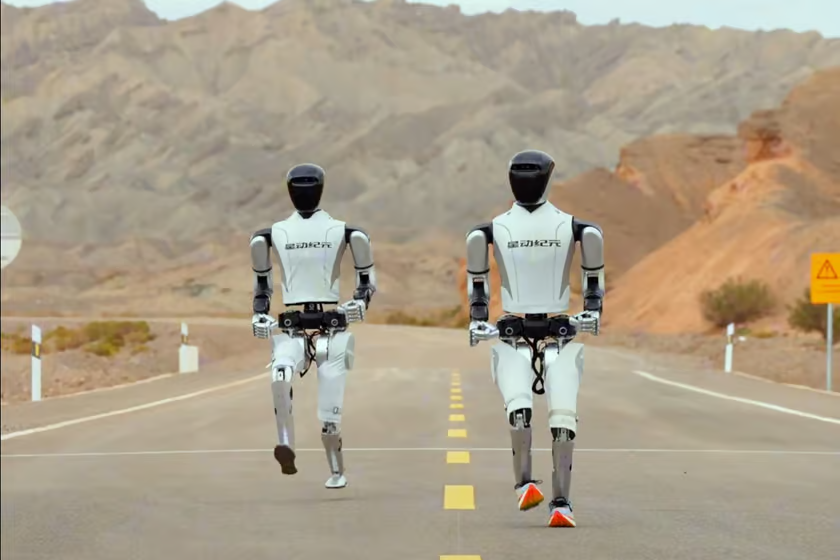 Androide con zapatillas deportivas vence a robot descalzo en una carrera en el Gobi Dos robots humanoides de la empresa Robot Era corren por una carretera en medio del desierto de Gobi. Ambos robots, con cuerpos blancos y negros, avanzan en línea recta, destacando sus piernas articuladas y torsos erguidos. El robot de la derecha lleva zapatillas naranjas, mientras que el de la izquierda corre descalzo. El paisaje detrás de ellos muestra montañas áridas y rocosas, creando un contraste entre la tecnología avanzada de los robots y el entorno natural desértico.