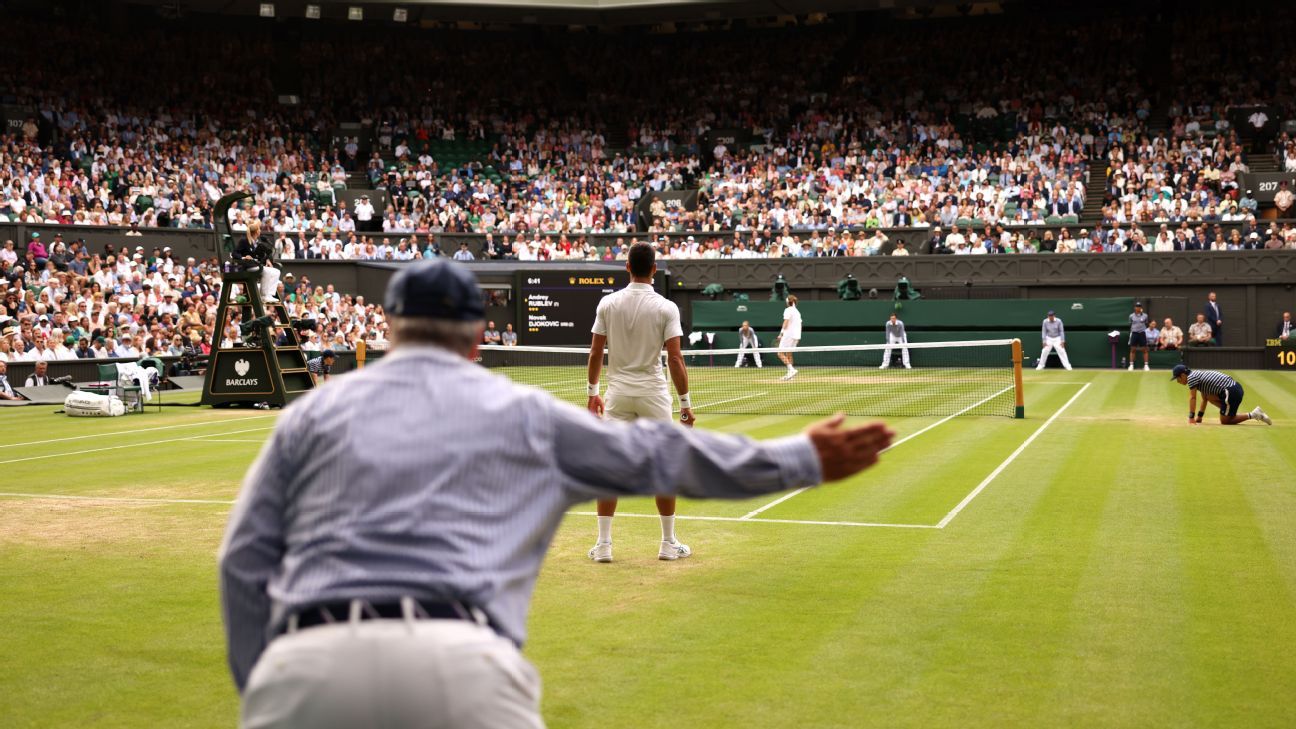 Wimbledon reemplazará a los jueces de línea con tecnología de inteligencia artificial a partir de 2025 Un árbitro de tenis levanta el brazo y señala con firmeza durante un partido en una cancha de césped, mientras uno de los jugadores espera de espaldas. La imagen muestra una multitud de espectadores en las gradas, con el juez de silla sentado en su plataforma en el centro de la escena. Los recogepelotas y jueces de línea están atentos en el campo, en lo que parece ser un torneo de gran importancia, probablemente Wimbledon, por el ambiente y el césped característico.