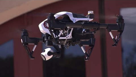 La policía de San Francisco estrena nuevo dron para combatir el crimen y aliviar la escasez de personal Primer plano de un dron profesional de alta tecnología en pleno vuelo, con una cámara incorporada y un diseño robusto en blanco y negro. El fondo muestra un edificio con ventanas grandes, ligeramente desenfocado.