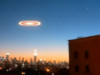 El auge de los vídeos falsos de drones sobre Nueva Jersey y el papel de la IA en su creación Una imagen que muestra un horizonte urbano al atardecer, con un cielo azul degradado hacia tonos anaranjados. En el centro del cielo, se ve un objeto circular brillante con un anillo luminoso, que parece suspendido sobre la ciudad. El paisaje incluye edificios y luces que destacan en la oscuridad creciente, con un rascacielos iluminado en el centro de la escena.