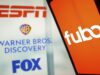 Disney adquiere FuboTV para consolidar su dominio en el streaming deportivo Logos de ESPN, Warner Bros. Discovery y Fox se muestran en un panel al fondo, mientras que en primer plano aparece la pantalla de un dispositivo móvil con el logotipo de Fubo en color blanco sobre un fondo naranja. La imagen representa la colaboración y acuerdos entre estas empresas en el contexto del streaming deportivo.