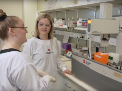 Avances revelan 293 nuevas variantes genéticas asociadas al trastorno de la depresión Dos investigadoras en un laboratorio científico, ambas usando batas blancas con el logo del QIMR Berghofer Medical Research Institute y gafas de seguridad. Una de ellas sostiene una conversación mientras están frente a un equipo automatizado de laboratorio de la marca Beckman Coulter, rodeado de tubos y cajas de reactivos. Al fondo, se observan estanterías con materiales y equipos de laboratorio.