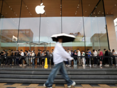 Apple elige a Alibaba para mejorar la inteligencia artificial en iPhones en China Una multitud de personas hace fila frente a una tienda de Apple en China, mientras un hombre con un paraguas camina frente a la escena en un día lluvioso. La fachada de vidrio refleja el entorno urbano y el icónico logo de Apple brilla sobre la entrada.