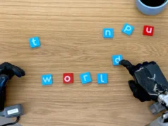 Google DeepMind lanza modelos de IA para un futuro más inteligente en robótica Dos manos robóticas manipulando pequeñas fichas con letras sobre una mesa de madera. Una de las manos está moviendo una ficha azul con la letra 'd'. En el centro de la imagen, las fichas forman la palabra 'world', con letras blancas sobre fondo azul y rojo. Otras fichas con letras dispersas están alrededor. En la esquina superior derecha, se ve parcialmente un objeto cilíndrico azul.