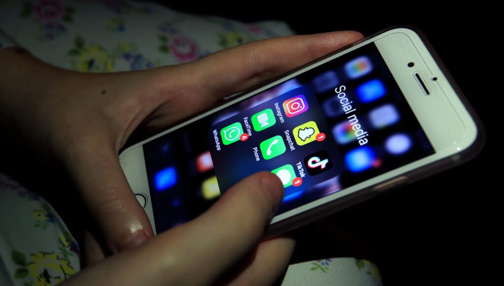 Persona sosteniendo un iPhone blanco con la pantalla encendida, mostrando una carpeta titulada "Social media" que contiene varias aplicaciones populares como Instagram, Snapchat, TikTok, WhatsApp, FaceTime y Phone. El entorno está oscuro y la luz de la pantalla ilumina ligeramente las manos del usuario.