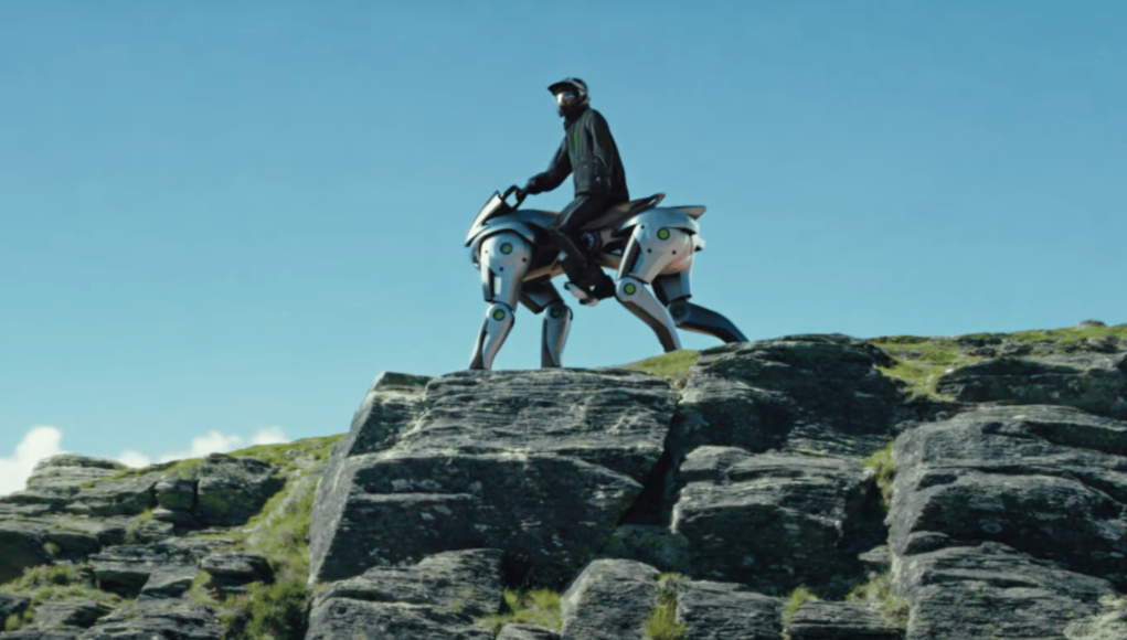 Persona montando un vehículo robótico cuadrúpedo de aspecto futurista sobre una formación rocosa al aire libre. El robot, con diseño metálico y articulaciones visibles, simula la forma de un animal de cuatro patas, mientras el piloto lleva casco y vestimenta de protección bajo un cielo despejado.