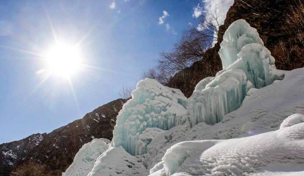 Formaciones de hielo y nieve en una ladera montañosa bajo un cielo despejado, con el sol brillando intensamente y árboles sin hojas al fondo, creando un contraste entre el clima frío y la luz solar.