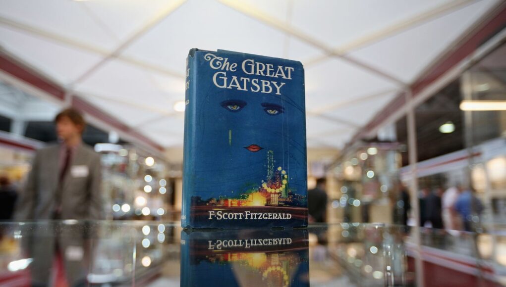 Un ejemplar de “The Great Gatsby” de F. Scott Fitzgerald, con su icónica portada azul mostrando un rostro con ojos y labios rojos sobre luces de la ciudad, se exhibe sobre una superficie de cristal en un espacio tipo feria o exposición, mientras se distingue la figura desenfocada de una persona al fondo.