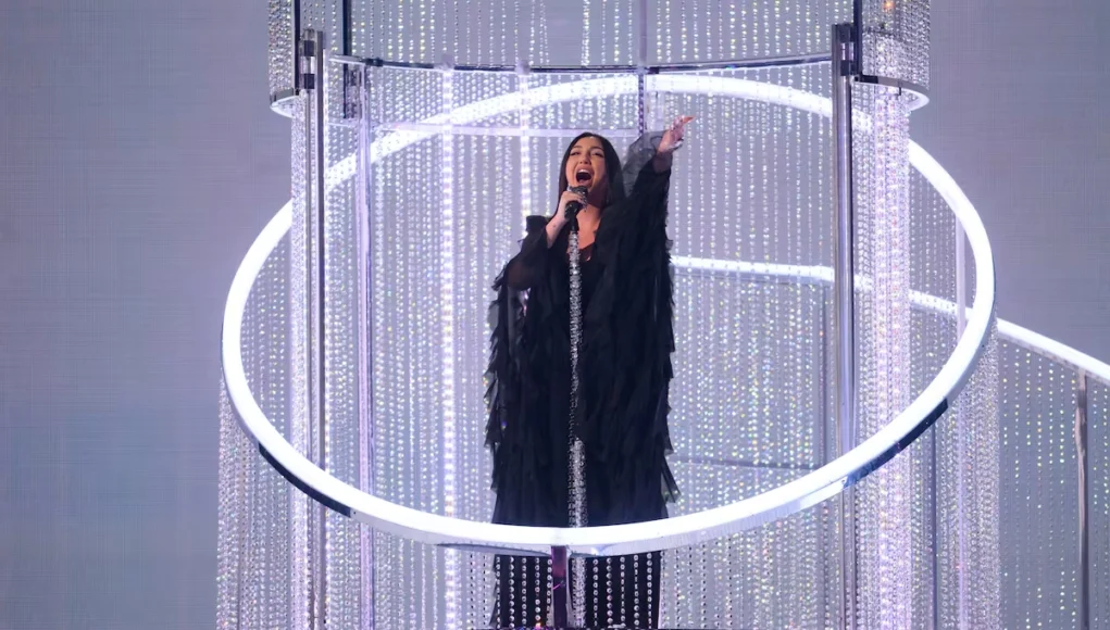 Polémica televoto Eurovisión Mujer cantando con un micrófono en una plataforma elevada rodeada de estructuras circulares y decoraciones brillantes, bajo una iluminación escénica intensa.