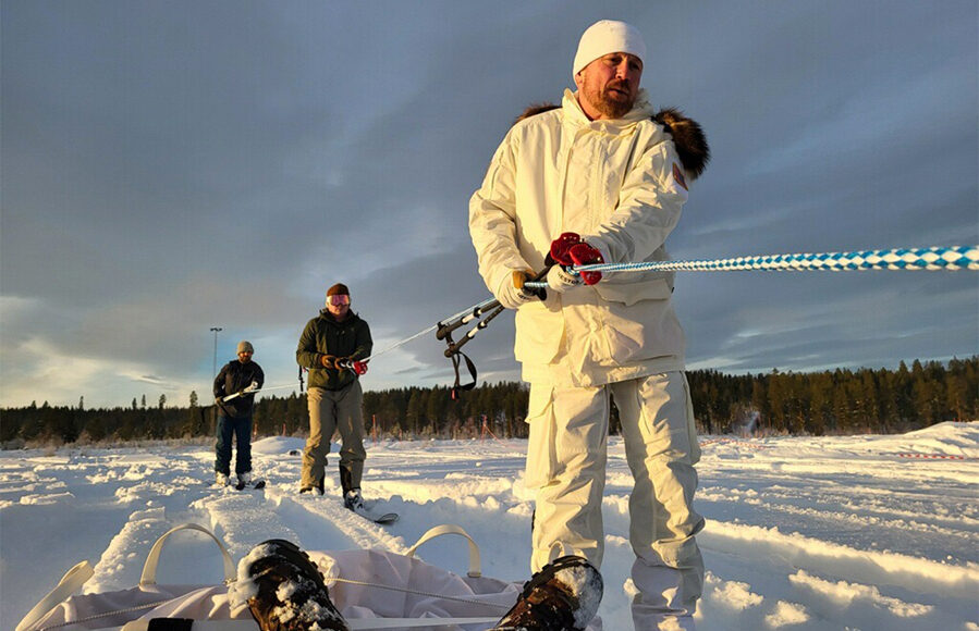 Tres personas arrastran un trineo en medio de un paisaje nevado; el hombre en primer plano viste ropa ártica blanca y sostiene una cuerda con esfuerzo.