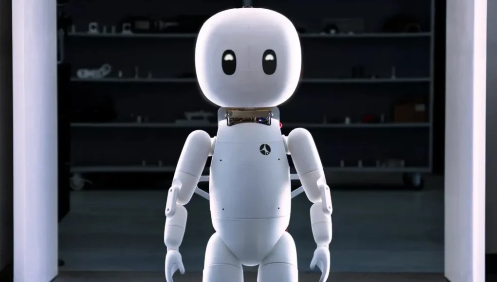 Robot humanoide pequeño de cuerpo blanco y diseño amigable, con cabeza redonda, ojos negros expresivos y extremidades articuladas, de pie sobre un suelo de baldosas en un entorno interior iluminado, frente a una zona oscura con estanterías al fondo.