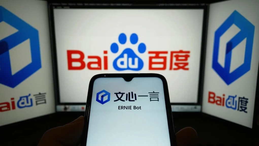Pantalla de un smartphone mostrando el logotipo y nombre del ERNIE Bot de Baidu en caracteres chinos y en inglés, con el logotipo hexagonal azul característico. Al fondo, varios monitores muestran el logo de Baidu en letras rojas y azules con la huella de pata.