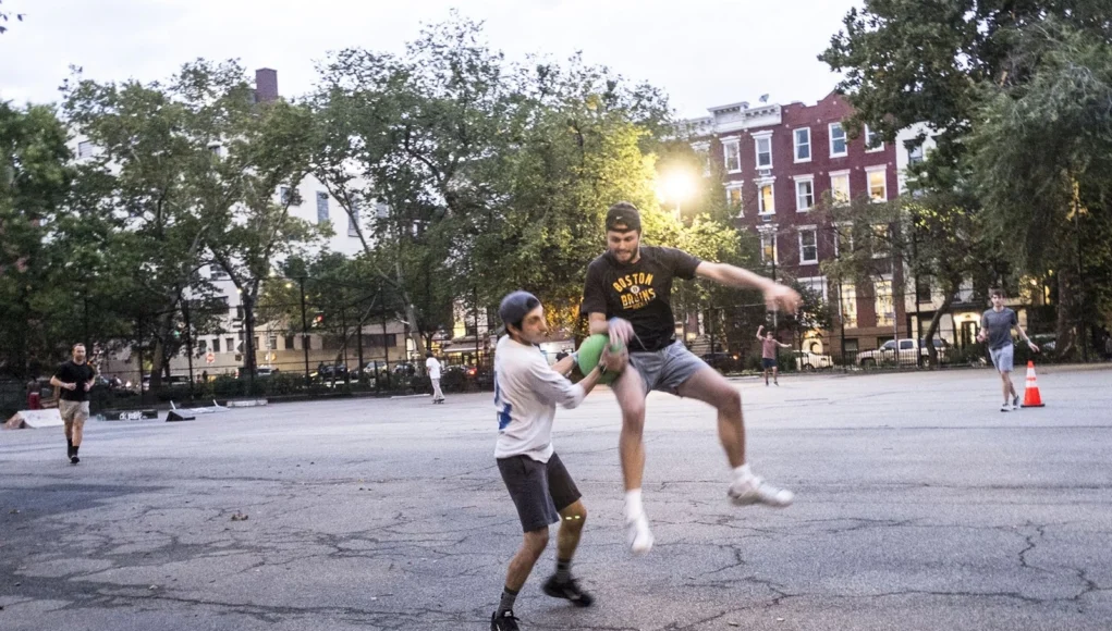 Dos hombres jugando kickball en una cancha urbana al atardecer; uno intenta alcanzar o bloquear al otro mientras sostiene una pelota verde, en una acción intensa y dinámica. Al fondo se observan más personas, árboles y edificios residenciales.