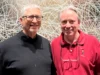 Bill Gates y Linus Torvalds se reúnen por primera vez en una cena histórica Dos hombres posan sonrientes frente a una pared con un fondo de líneas entrelazadas; uno lleva gafas y un suéter negro, y el otro una camiseta tipo polo roja con un dibujo bordado de un personaje animado.