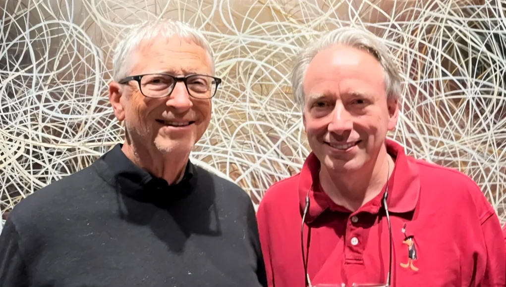Dos hombres posan sonrientes frente a una pared con un fondo de líneas entrelazadas; uno lleva gafas y un suéter negro, y el otro una camiseta tipo polo roja con un dibujo bordado de un personaje animado.