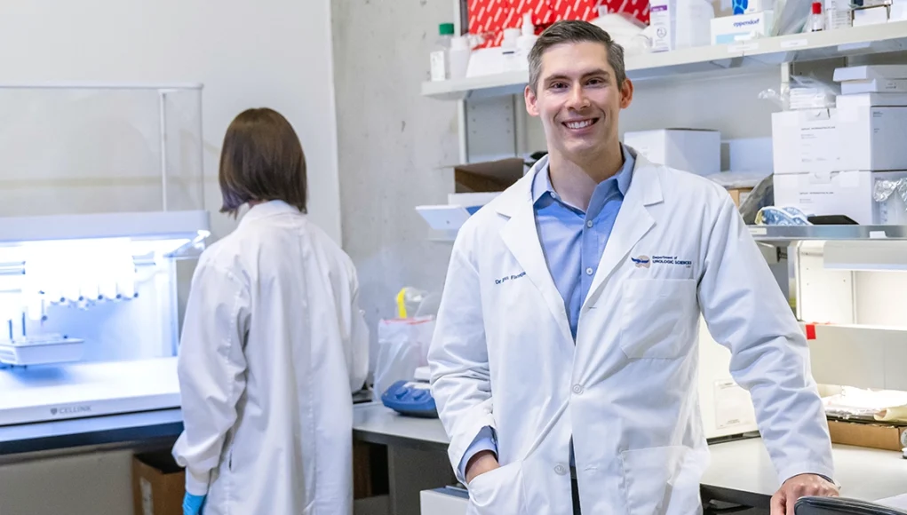 Dos investigadores en un laboratorio moderno; uno de ellos, un hombre con bata blanca y camisa azul, sonríe mientras mira a cámara, mientras que otra persona trabaja de espaldas frente a un equipo científico.