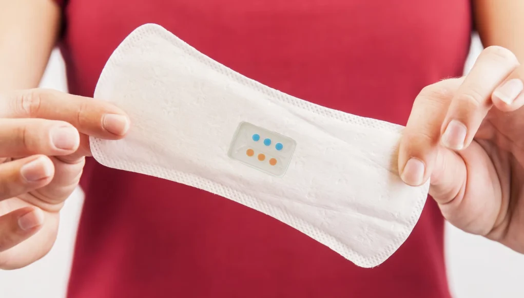 Primer plano de una persona sosteniendo una compresa blanca con un sensor integrado que muestra indicadores de colores, posiblemente diseñado para monitorear la salud o detectar infecciones.