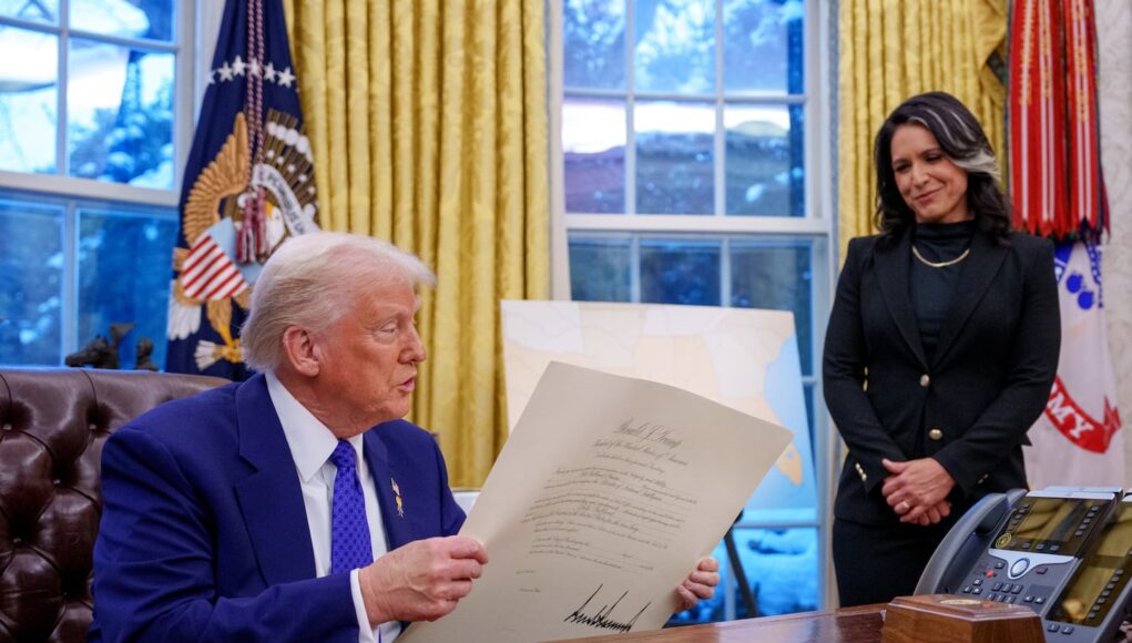 Un hombre de cabello rubio, vestido con traje azul, sostiene un documento oficial sentado en un escritorio, mientras una mujer vestida de negro lo observa de pie con una expresión de respeto. Están en una oficina con cortinas doradas, banderas oficiales y teléfonos sobre el escritorio.