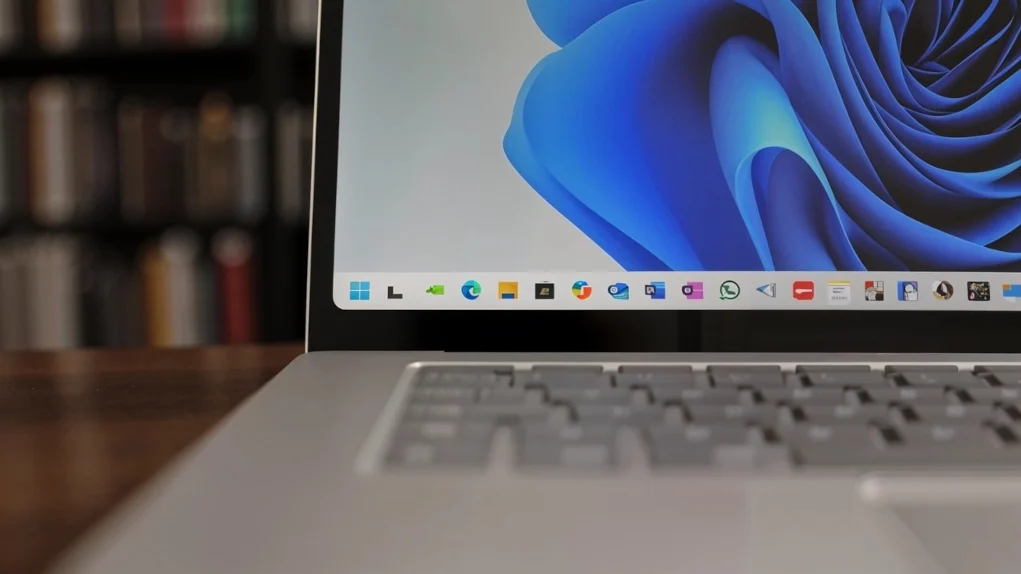 Primer plano del borde inferior de la pantalla de un portátil con Windows 11, mostrando el menú de aplicaciones ancladas en la barra de tareas. En el fondo, se aprecian estanterías desenfocadas llenas de libros.