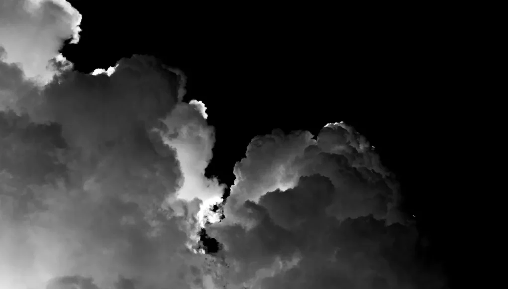 Nubes densas e iluminadas parcialmente desde atrás, en una fotografía en blanco y negro con un cielo oscuro que genera un contraste dramático.