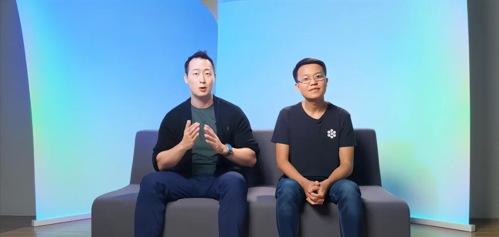 Dos hombres sentados en un sofá frente a un fondo degradado azul claro. El hombre a la izquierda gesticula mientras habla y lleva un reloj azul, camiseta verde y chaqueta negra. El hombre a la derecha, con gafas y camiseta negra con un logotipo blanco, permanece tranquilo con las manos entrelazadas.