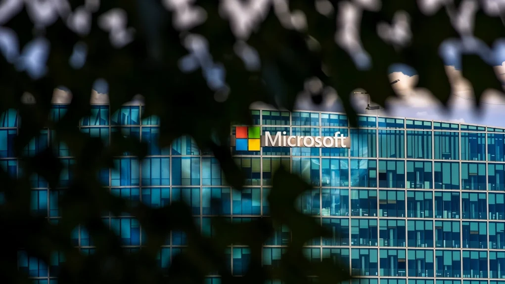 Vista parcial de la sede de Microsoft a través de hojas desenfocadas en primer plano, mostrando el logo de la compañía en un edificio moderno de cristal azul.
