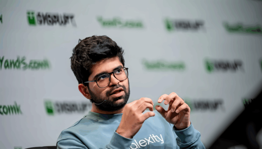 Hombre joven con gafas y barba hablando en un evento tecnológico, con un micrófono de diadema y gesticulando con las manos. Lleva una sudadera azul con la palabra "Alexity" impresa, y al fondo se distingue el logo de "Disrupt" repetido en el panel del escenario.