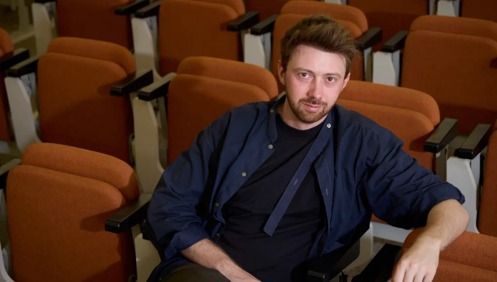 Hombre con expresión seria sentado en una butaca de un auditorio vacío, con filas de asientos naranjas a su alrededor. Viste una camisa azul abierta sobre una camiseta negra y mira directamente a la cámara.