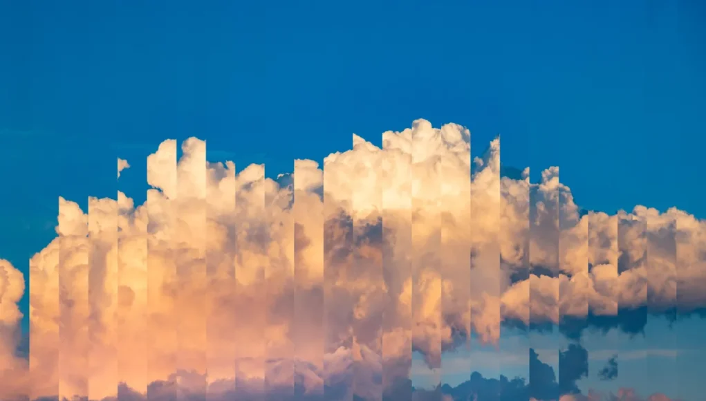 Nubes algodonosas de tonos cálidos sobre un cielo azul, con un efecto visual de repetición o distorsión digital que divide la imagen en columnas verticales, creando una apariencia fragmentada y artística del cielo.