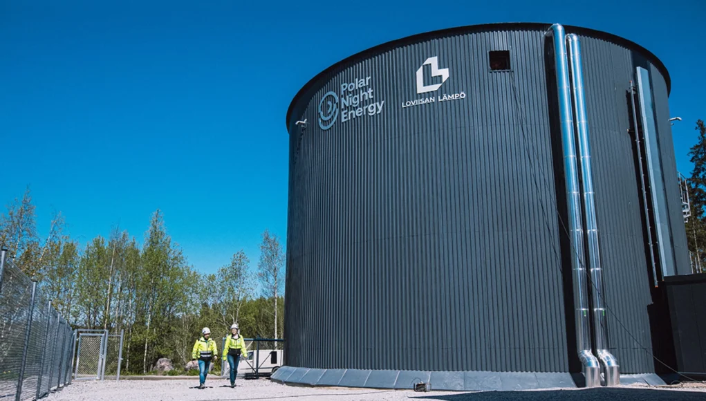Dos personas con chalecos reflectantes y cascos caminan junto a un gran silo cilíndrico gris oscuro de la empresa Polar Night Energy y Lovisan Lämpö, bajo un cielo despejado, rodeado de árboles y estructuras industriales.