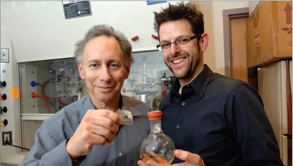 Dos personas en un laboratorio posan para la cámara. La persona a la izquierda sostiene un pequeño cuadrado translúcido que parece un material experimental, mientras que la persona a la derecha sostiene un matraz esférico con tapón naranja. Al fondo se observan equipos y mobiliario de laboratorio, incluyendo una campana extractora.