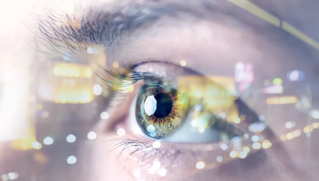 Primer plano de un ojo humano con reflejos brillantes de luces de ciudad superpuestos, simbolizando tecnología, percepción aumentada o visión futurista. El iris muestra detalles en tonos verdes y dorados, con enfoque en la pupila y las pestañas.