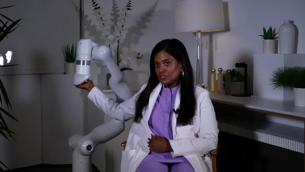 Una mujer con bata blanca y blusa morada está sentada mientras sostiene el extremo de un brazo robótico blanco. El entorno es moderno y elegante, con plantas, lámparas y decoración de oficina al fondo.