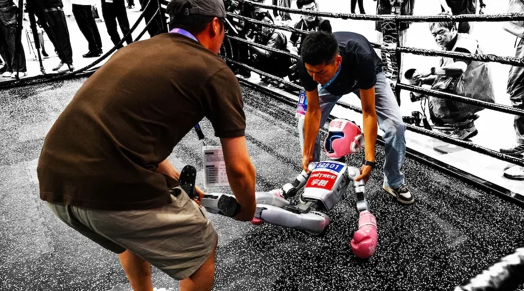 Un pequeño robot humanoide con guantes de boxeo siendo asistido tras caer en lo que parece un ring de combate, mientras varias personas lo observan y fotografían desde el exterior. La escena, capturada con un efecto visual que resalta en color solo al robot y a sus cuidadores, refleja de forma simbólica los desafíos actuales en el desarrollo de la robótica, donde el espectáculo y la innovación se cruzan con la fragilidad de la tecnología en evolución.
