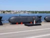 La Royal Navy controla un submarino robótico desde 16,000 kilómetros de distancia Un prototipo de submarino no tripulado (UUV) presentado oficialmente en un acto militar, con un oficial de la marina británica dando un discurso frente a la unidad. El vehículo, de diseño compacto y aerodinámico, está expuesto junto al mar sobre soportes naranjas, destacando su uso potencial en misiones submarinas de vigilancia, reconocimiento o guerra electrónica sin necesidad de tripulación humana.