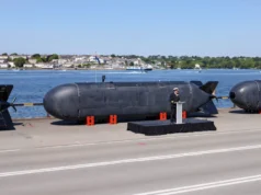 La Royal Navy controla un submarino robótico desde 16,000 kilómetros de distancia Un prototipo de submarino no tripulado (UUV) presentado oficialmente en un acto militar, con un oficial de la marina británica dando un discurso frente a la unidad. El vehículo, de diseño compacto y aerodinámico, está expuesto junto al mar sobre soportes naranjas, destacando su uso potencial en misiones submarinas de vigilancia, reconocimiento o guerra electrónica sin necesidad de tripulación humana.