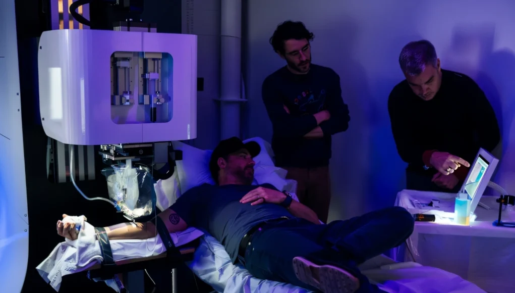 Persona recostada en una camilla recibiendo un tatuaje realizado por una máquina controlada por inteligencia artificial, mientras dos hombres observan y supervisan el proceso en un entorno iluminado con luces azules.