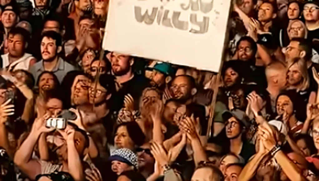 Multitud de personas en un concierto o evento masivo, muchas de ellas aplaudiendo, grabando con sus teléfonos móviles o cámaras. En el centro se destaca un cartel levantado sobre la multitud que dice “WILLY”, posiblemente en referencia a un artista. La imagen transmite entusiasmo y energía colectiva.