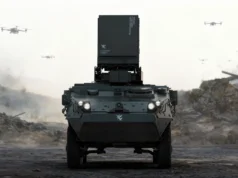 Arma de microondas Epirus Leonidas derriba 49 drones con un solo disparo Vehículo militar blindado con sistema montado de defensa contra drones, avanzando en un campo de batalla con varios drones volando en el cielo y humo en el fondo.