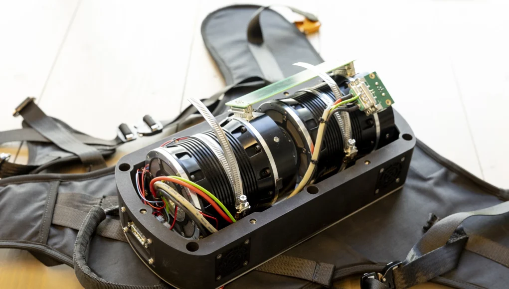 Dispositivo mecánico y electrónico montado en una estructura de arnés con correas, que muestra cables de colores, ventiladores y componentes metálicos.