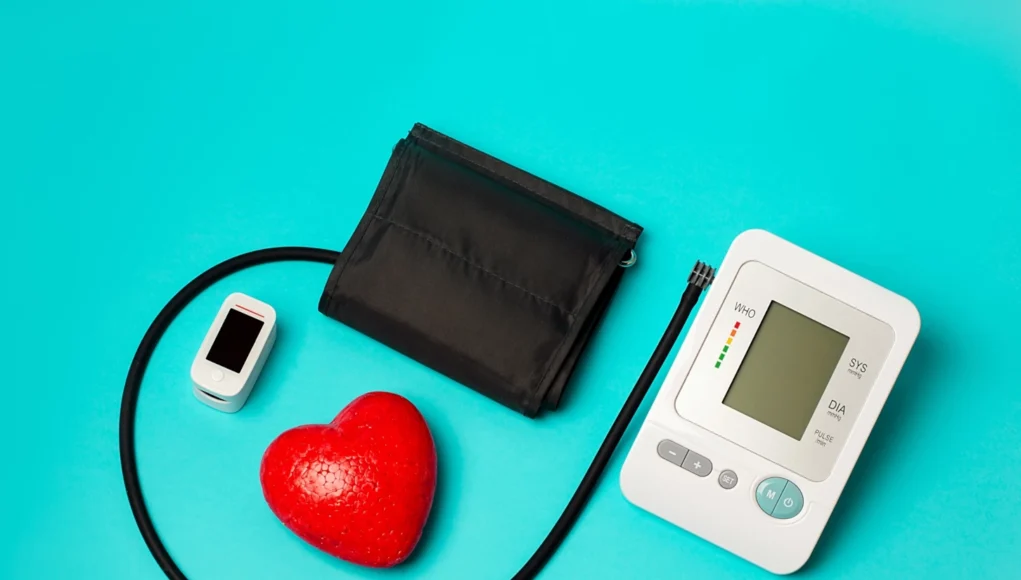 Control Hipertension Persistente Equipo médico sobre fondo azul: un tensiómetro digital con brazalete negro, un oxímetro de pulso blanco y un corazón rojo decorativo, simbolizando el monitoreo de la salud cardiovascular.