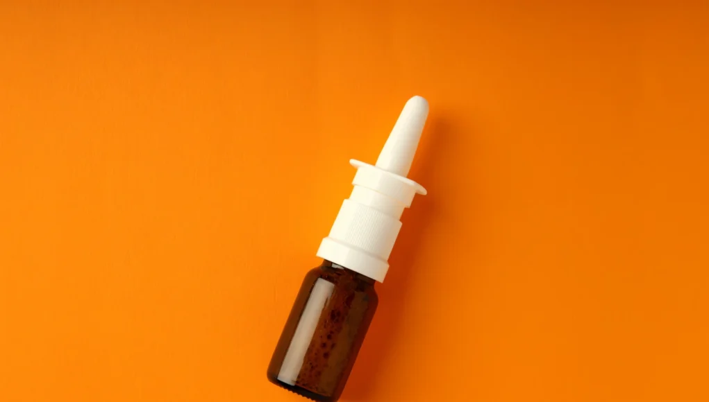 Spray Diurético Frasco marrón pequeño con aplicador en spray nasal blanco, colocado sobre un fondo naranja.