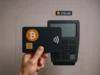 El auge de las tarjetas cripto revoluciona los pagos digitales Una mano sostiene una tarjeta de color negro con el logotipo de Bitcoin y el símbolo de pago sin contacto, frente a un terminal de pago electrónico de color gris. En la parte superior se ve el nombre “bitcoin” acompañado de su logotipo.
