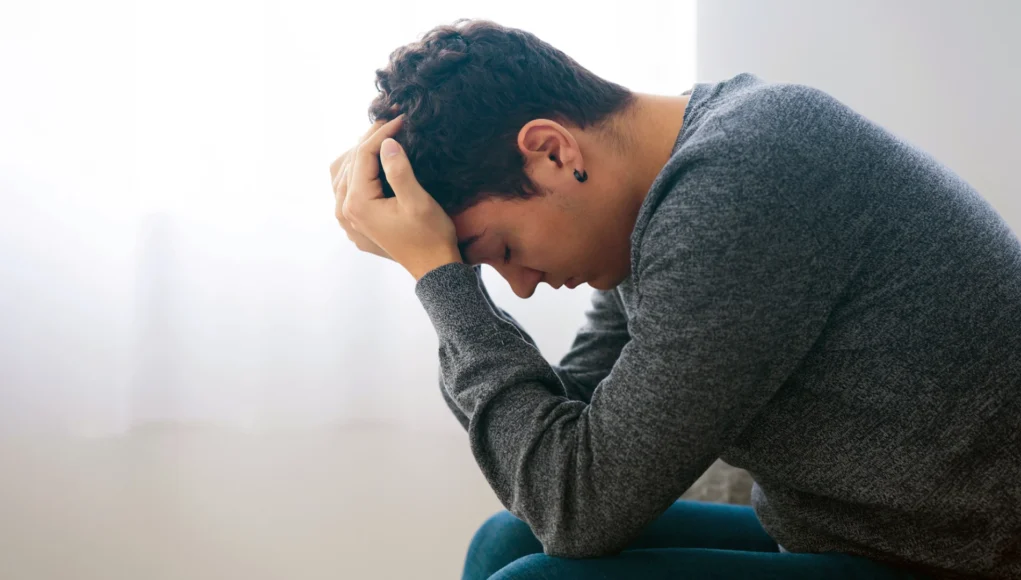 Joven sentado con la cabeza inclinada hacia adelante y las manos sobre el cabello, mostrando una expresión de angustia o preocupación.