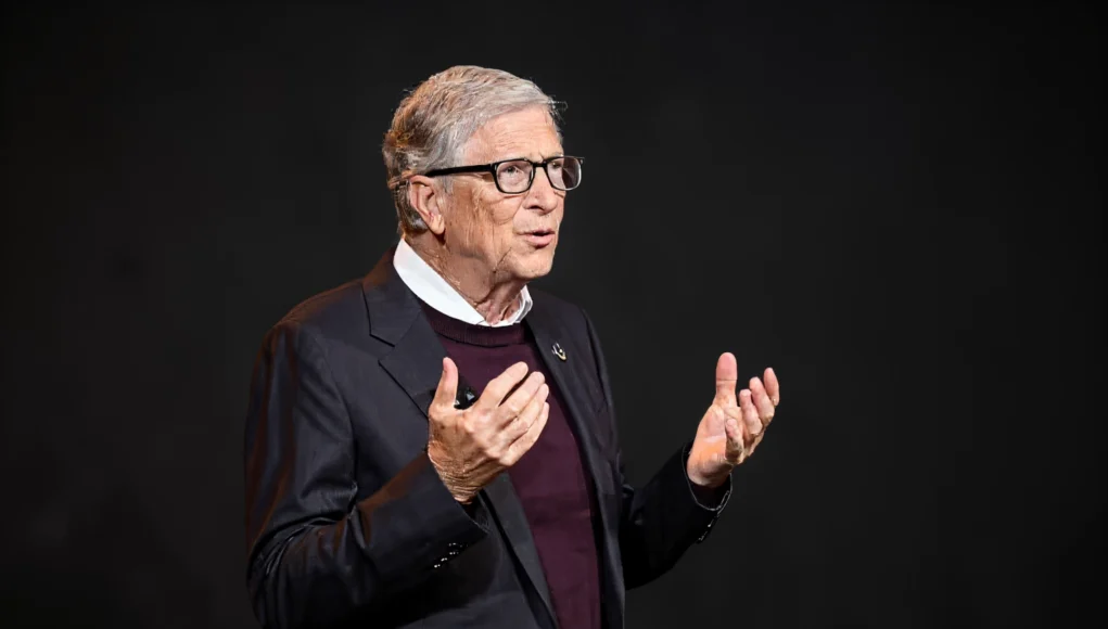 Hombre mayor con gafas y traje oscuro hablando en un escenario, gesticulando con las manos frente a un fondo negro.