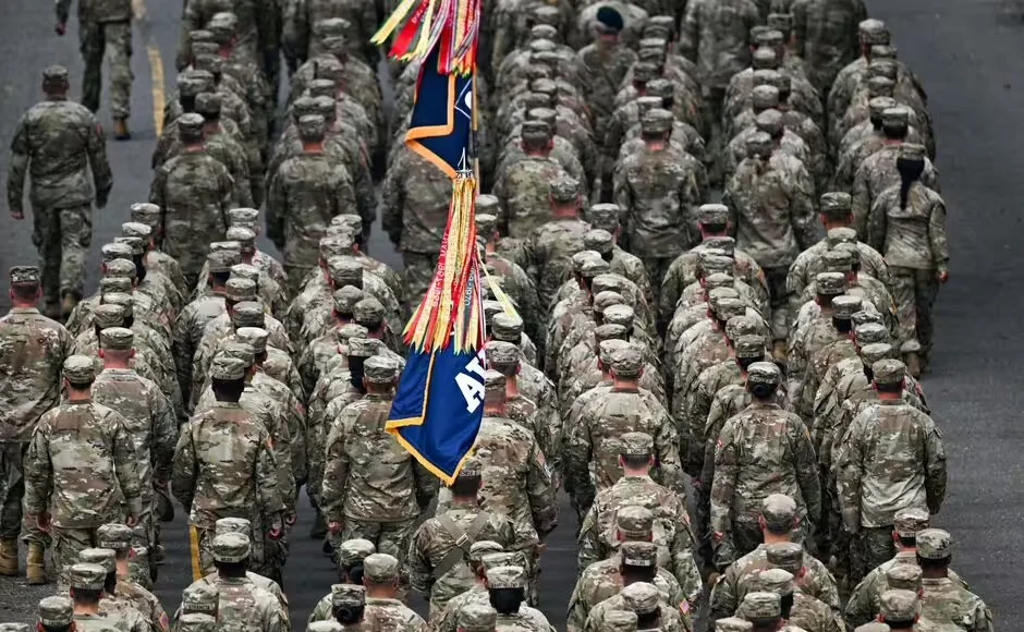 Grupo de soldados uniformados marchando en formación, vistos desde atrás, con estandartes militares decorados con cintas de colores en el centro de la fila.