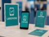 La Free Software Foundation lanza el proyecto Librephone para liberar los teléfonos móviles Stand promocional del proyecto LibrePhone, con un teléfono móvil en exposición acompañado de carteles y folletos en color turquesa que muestran el logotipo de un smartphone con una marca de verificación.