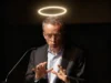 Patrick Gelsinger pretende impulsar una IA cristiana con el objetivo de acelerar la llegada de Cristo Hombre mayor con traje y gafas hablando en un atril con micrófono, bajo una luz circular que parece un halo iluminado sobre su cabeza.
